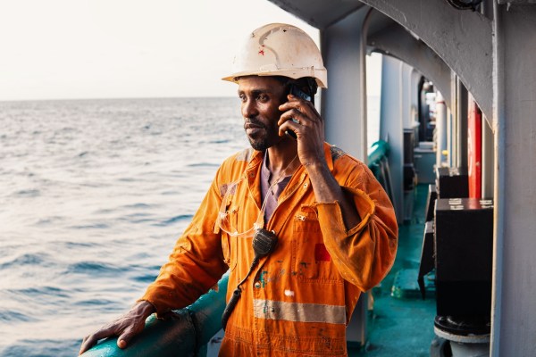 A sailor on the deck of a ship, talking on a phone and staring at the horizon.